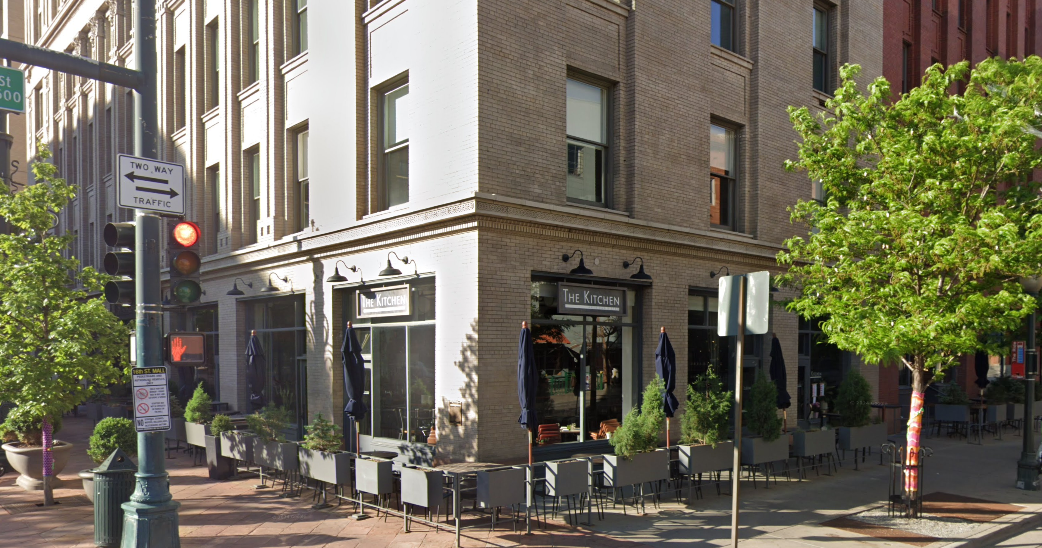 The image shows a street corner with a brick building housing a restaurant called "The Kitchen." There are large windows and outdoor seating with tables and chairs. A traffic light is visible, showing a red light, along with a "Two Way Traffic" sign. There are trees with green leaves along the sidewalk, and a pedestrian crossing signal is also present.